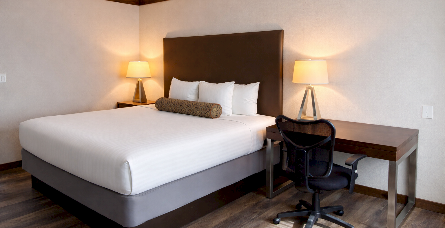 A neatly made hotel room with a large bed, two lamps, a desk, and a chair under a dark headboard.