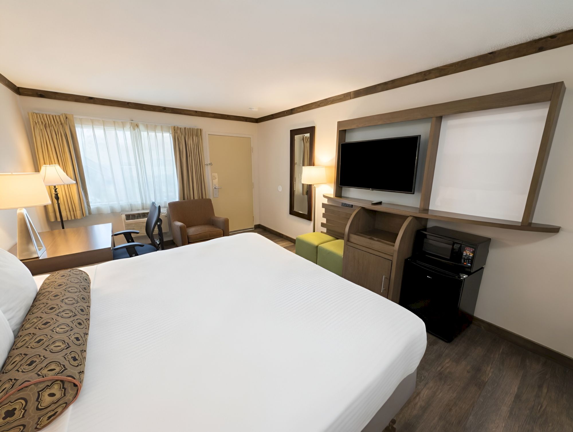 A modern hotel room with a large bed, armchairs, window curtains, and a wall-mounted flat-screen TV.