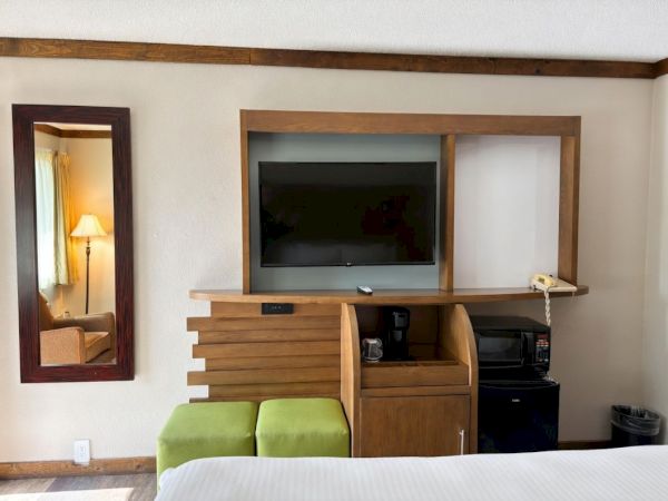 A hotel room with a wall-mounted TV in a wooden cabinet, coffee maker and microwave below, plus green seating and a lamp-lit mirror.
