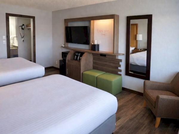 A modern hotel room with two beds, a TV, desk, green ottoman, lamp, and mirrors.