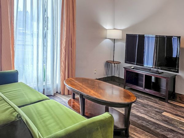 A bright living room with a green sofa, oval wooden coffee table, TV stand, and floor lamp beside large windows.