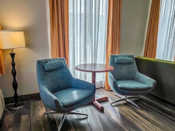 A cozy hotel room seating area with two blue swivel chairs, a round table, and tall curtains.