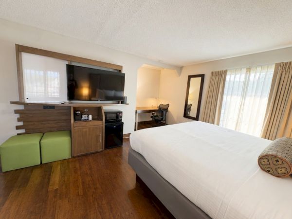 A modern hotel room with a large bed, flat-screen TV on a cabinet, office desk, green bench, and bright curtains.
