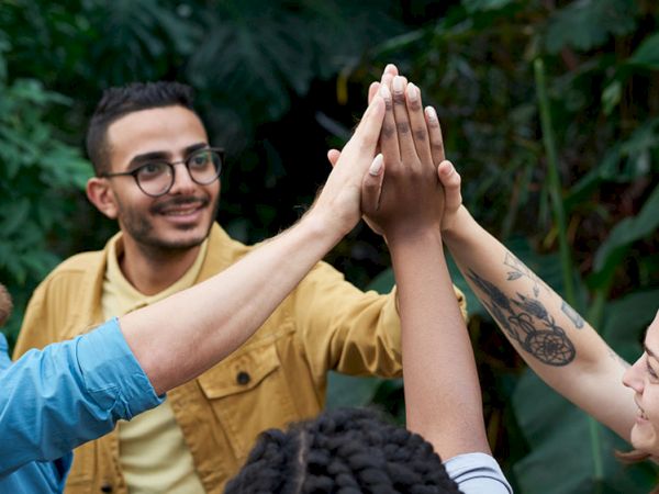 A man and woman give a friend a high-five outdoors in greenery, smiling and celebrating teamwork.