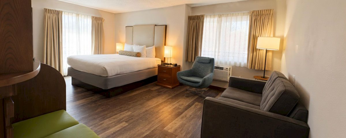 A cozy hotel bedroom with a queen bed, dark sofa, chairs, lamps, and large windows with curtains, all neatly arranged.