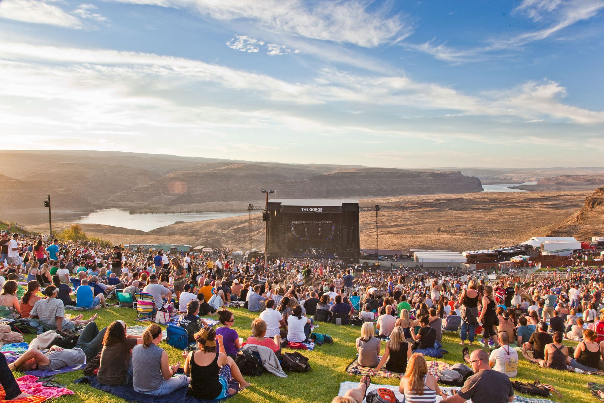 A crowd sits on grass for a live music show at an outdoor canyon amphitheater near a winding river.