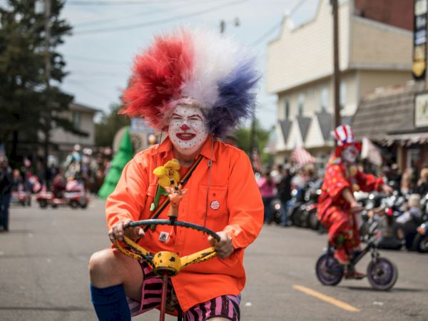 A clown in an orange suit with a red-and-blue wig and painted face rides a decorated tricycle in a street parade.