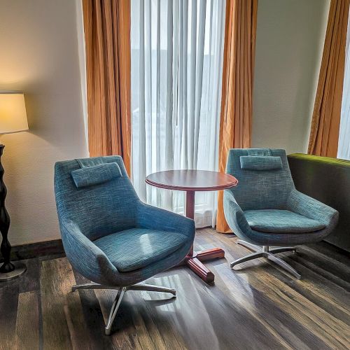A cozy living room with two blue armchairs, a small round table, a floor lamp, and large curtains behind.