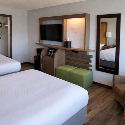 A hotel room with two beds, a flat-screen TV on a console, a green ottoman, a chair, and doorways to the bathroom and another room.
