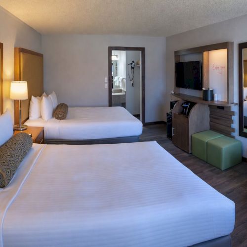 A modern hotel room with two neatly made beds, bedside lamps, a TV, a desk, and a mirror, lit by warm overhead light.