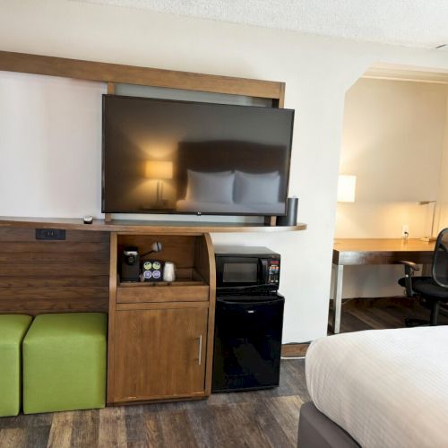 A hotel room setup with a flat-screen TV on a wooden cabinet, mini-fridge, microwave, desk, and bed.