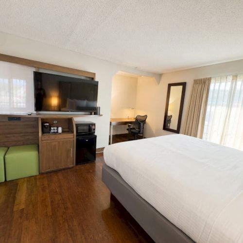Modern hotel room: bed, wall TV on wooden cabinet, desk chair, and window curtains.