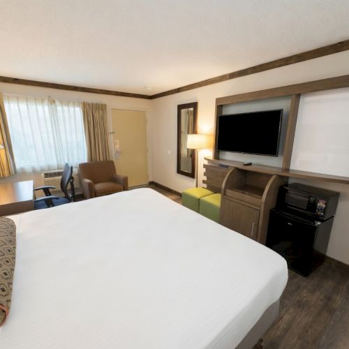 A hotel room with a large bed, TV on a console, lamps, curtains, and a sitting area with chairs and a green ottoman.