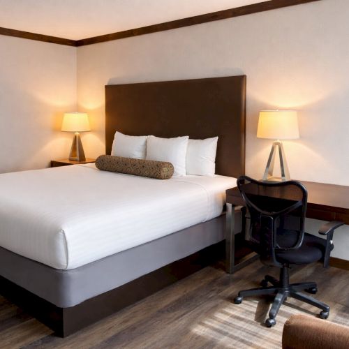 A neatly made hotel-style bedroom with a large bed, two bedside lamps, and a desk with a black office chair.