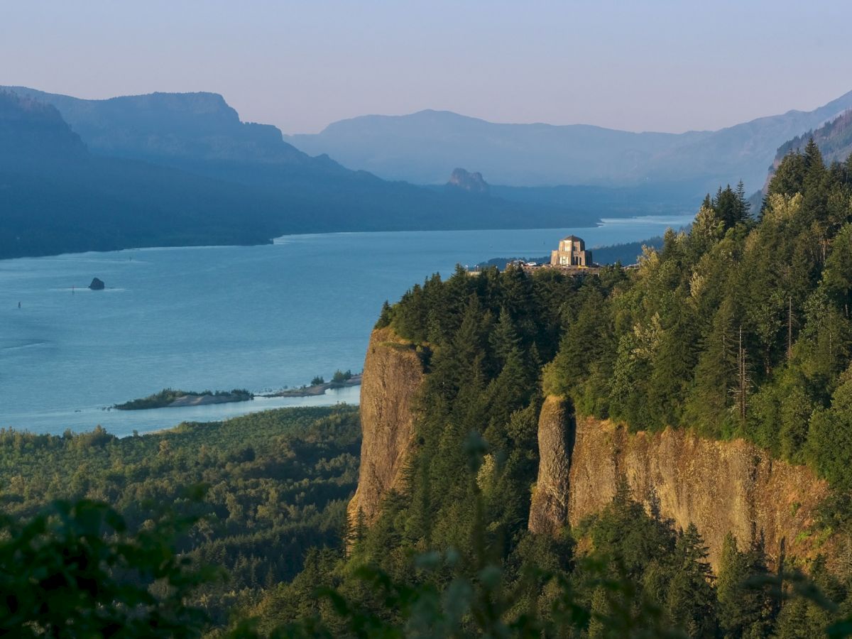 Scenic mountain valley with a forested cliff overlooking a calm lake and a small chapel-like building near the shore.