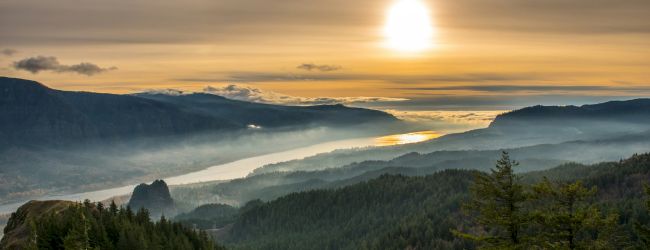 A golden sunrise over a misty valley with winding river-like water, layered hills, and dense evergreen forests.