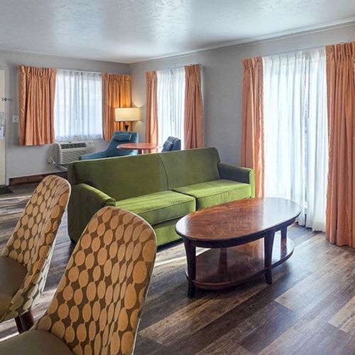 A cozy living room with a green sofa, patterned armchairs, round coffee table, and large windows with brown curtains.