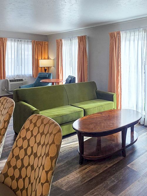 A cozy living room with a green sofa, patterned armchairs, round coffee table, and large windows with brown curtains.