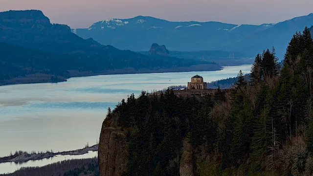 A wide river-lake landscape with mountains and a small dome-like monument on a cliff overlooking the water at dusk.