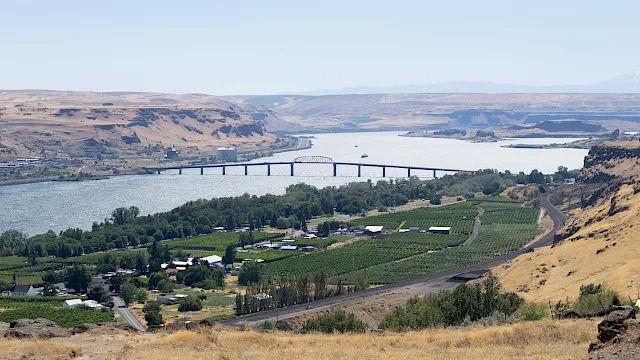 A long bridge spans a wide river across dry hills, with farmland and houses along the banks beneath a clear sky.