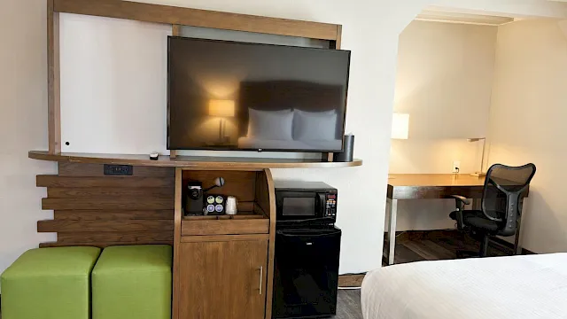 A hotel room with a flat-screen TV on a wooden cabinet, mini fridge, microwave/coffee items, and a desk and chair.