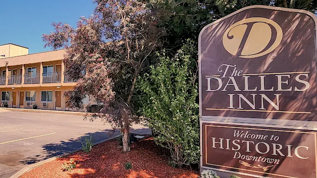 A roadside sign reads &ldquo;The Dalles Inn&rdquo; and &ldquo;Welcome to Historic Downtown,&rdquo; beside hotel buildings and trees in a parking lot.